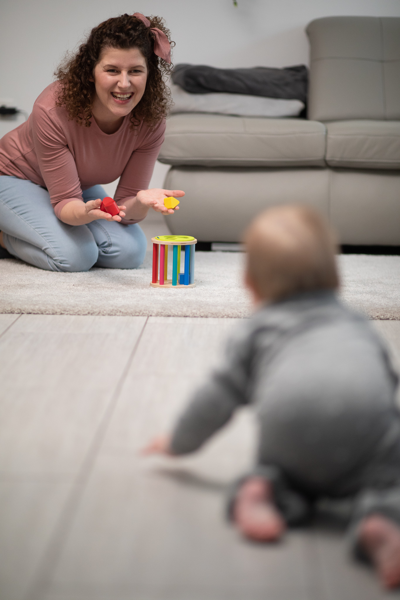 Mama Franzi im Hintergrund kniend auf dem Boden auf einem Teppichmit roten und gelben Bausteinen in den Händen, vor ihr ein Holzsteckspiel, Baby Mini im Vordergrund krabbelt auf Mama zu