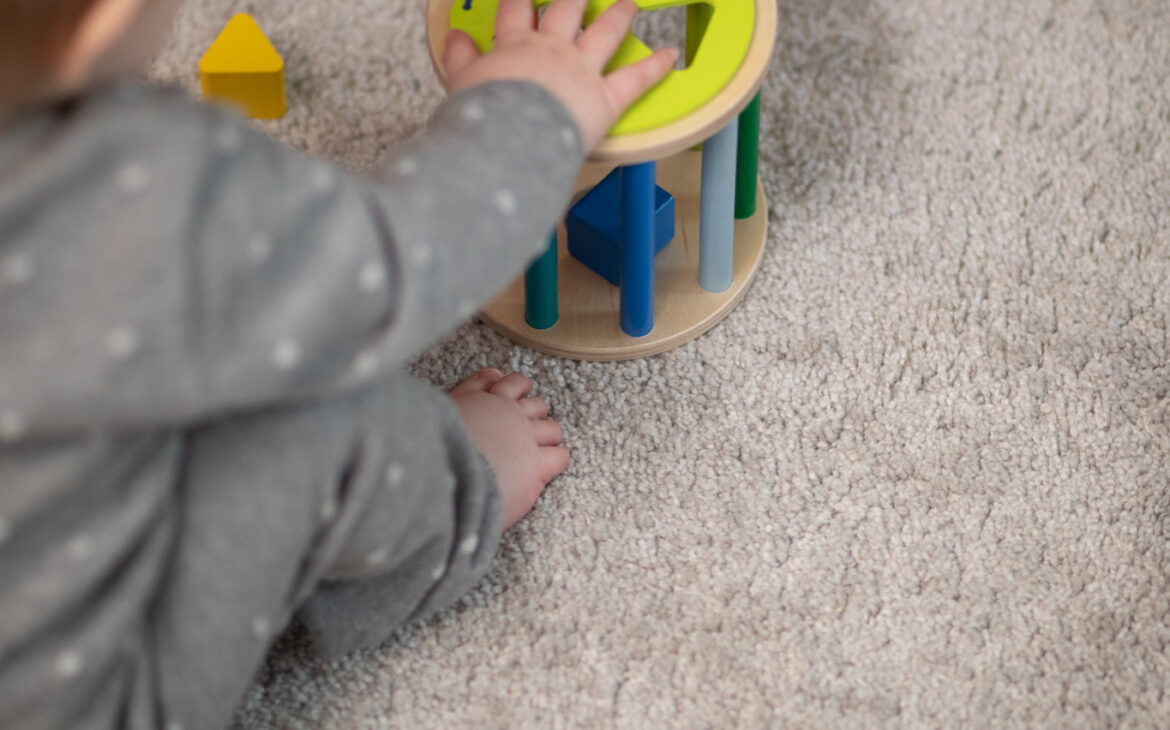 Baby in grauem Overall sitzend im Vordergrund greift mit der rechten Hand ein Holzsteckspiel