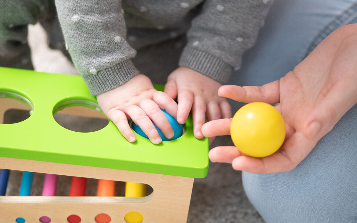 Zwei kleine Kinderhände, die eine blaue Holzkugel in ein Holzsteckspiel mit grünem Deckel und bunten Stäben, stecken. Eine größere Hand, die der Mama, reicht dem Kind eine gelbe Holzkugel an
