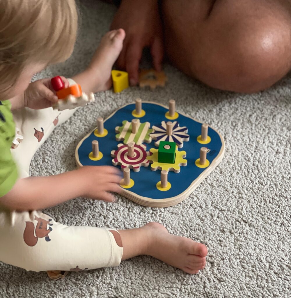 Seitliche Vogelperspektive von rechts oben auf die Szene Papa und Kleinkind spielen zusammen am Boden auf dem beigefarbenen Teppich. Vom Papa ist am oberen rechten Bildrand nur ein kleiner Teil des Knies zu sehen. Mini sitzt im Bild links, vor ihr der Selecta Kurbelspaß. Mit ihrer rechten Hand greift sie in Richtung des blauen Spielbretts und in der linken Hand hält sie ein hölzernes Zahnrad fest, sowie einen roten und orangefarbenen Holzbaustein.