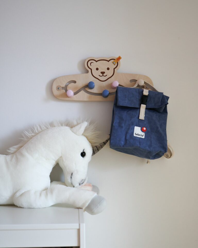 Eine Kindergarderobe aus Holz von Steiff hängt an einer weißen Wand. Die Aufhängelemente sind von links nach rechts rosa, blau, blau, rosa und holzfarben. Am ganz rechten Element ist ein dunkelblauer Rucksack aufgehängt, der die Aufschrift Selecta trägt. Im Bild links unten sieht man ein weißes Plüscheinhorn, dass auf einem Schrank liegt.